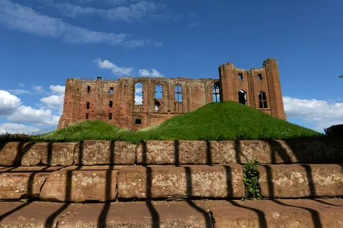 The image shows an architectural photograph of Kenilworth Castle in the town of Kenilworth, Warwickshire. Captured in the late afternoon during spring, the photograph features the historic ruins of Kenilworth Castle, an English Heritage site, as seen from behind a grate with shadows cast across the stone steps in the foreground. The castle, built of red sandstone, stands prominently against a blue sky with scattered clouds and is surrounded by lush green mounds and well-kept grass typical of the spring season. This English Heritage castle is a significant landmark, reflecting centuries of architectural history in Kenilworth.